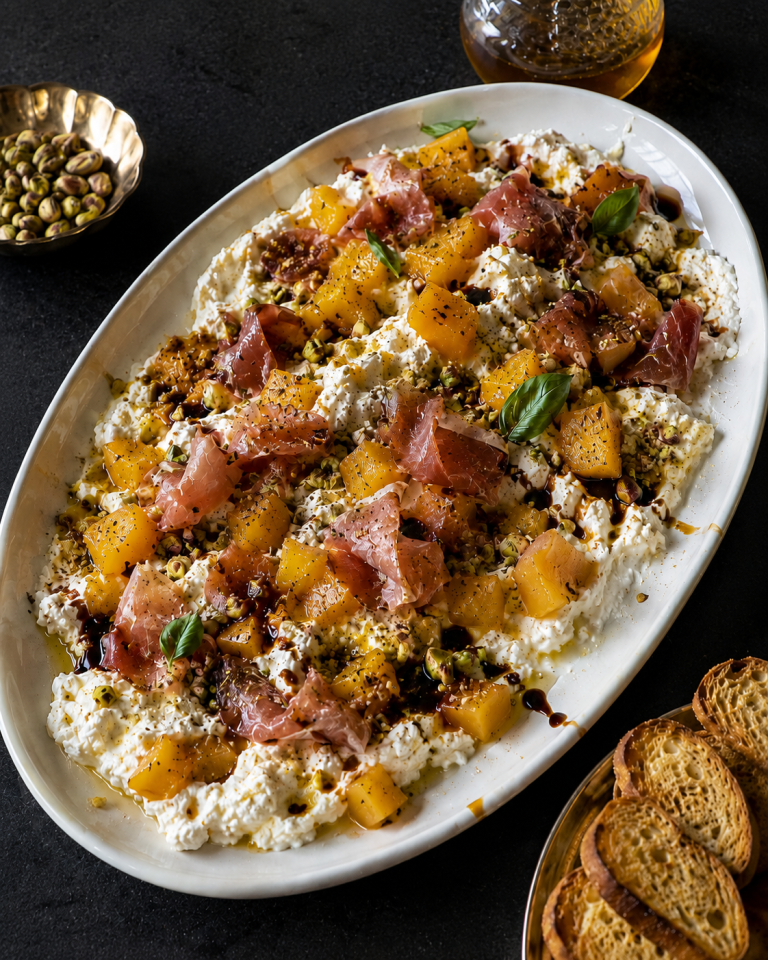 Mango prosciutto burrata dip with hot honey, basil, pistachios and crostini in a white oval dish