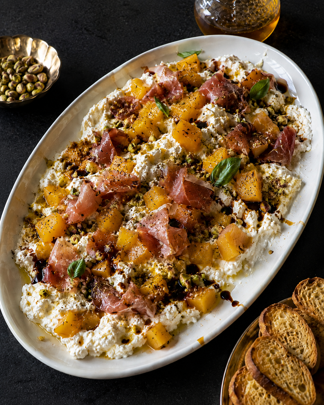 Mango prosciutto burrata dip with hot honey, basil, pistachios and crostini in a white oval dish