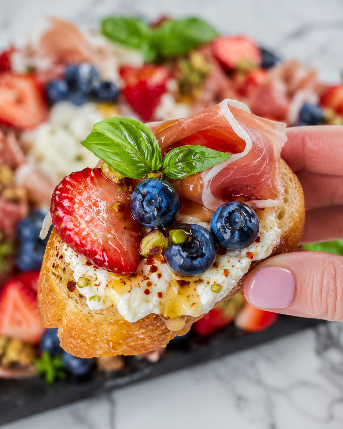 Close-up of a hand sprinkling Firecracker Party Dust over a red white and blue burrata appetizer board with burrata, strawberries, blueberries, prosciutto, crostini, basil, hot honey, and pistachios on a black tray.