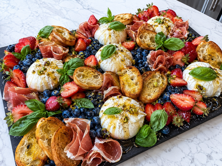 Huge red white and blue burrata appetizer board with strawberries, blueberries, prosciutto, crostini, basil, hot honey, and pistachios on a black tray.