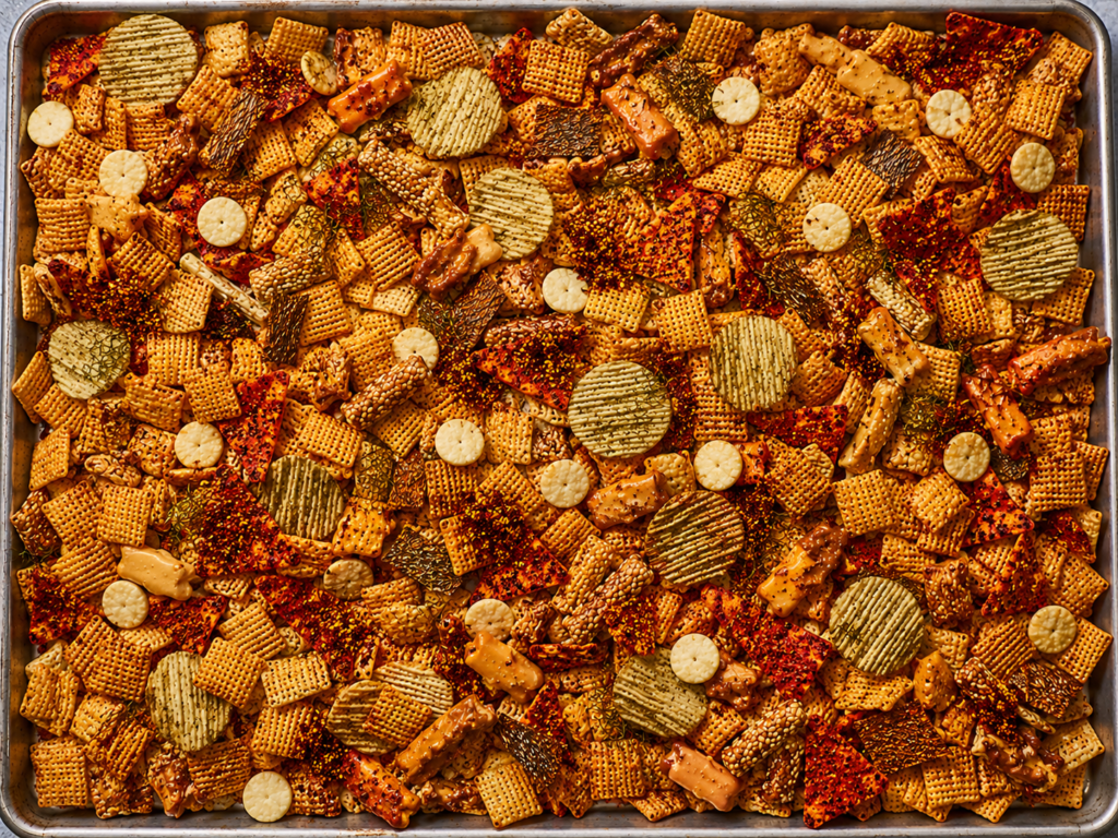 Cooked Bloody Mary dill pickle Chex Mix spread across a parchment-lined baking sheet with crisp cereal, pickle chips, oyster crackers, cheese-filled Combos, rye chips, and red cornmeal clusters.
